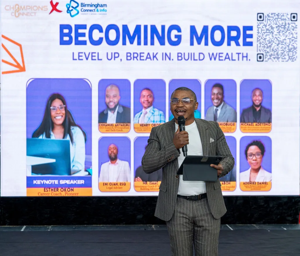 A host speaking into a microphone in front of the Becoming More screen showing the speaker line-up