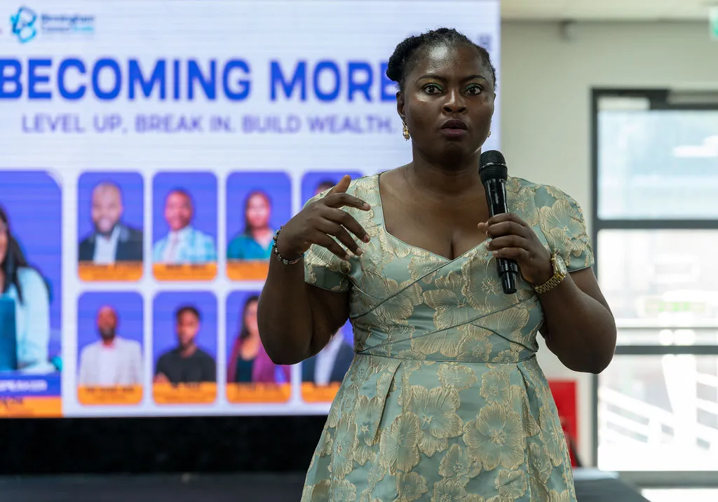 A speaker in a green dress gesturing as she presents beside the Becoming More screen