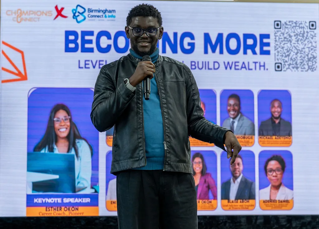 A speaker in a leather jacket smiling as he presents in front of the stage screen