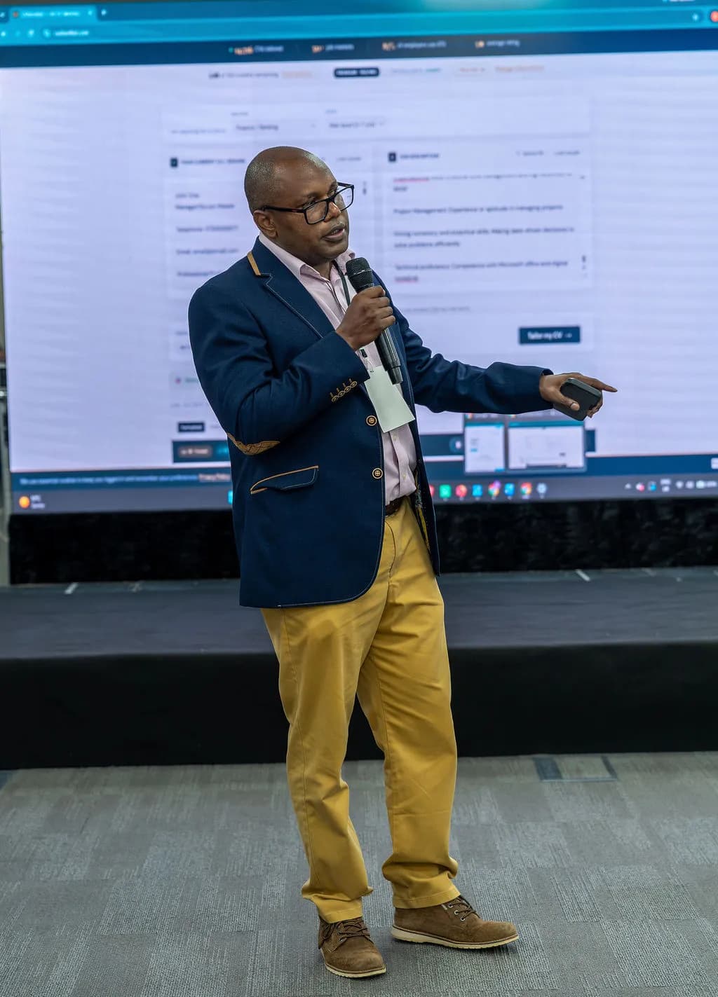A speaker in a navy blazer and mustard trousers pointing at a presentation on the big screen